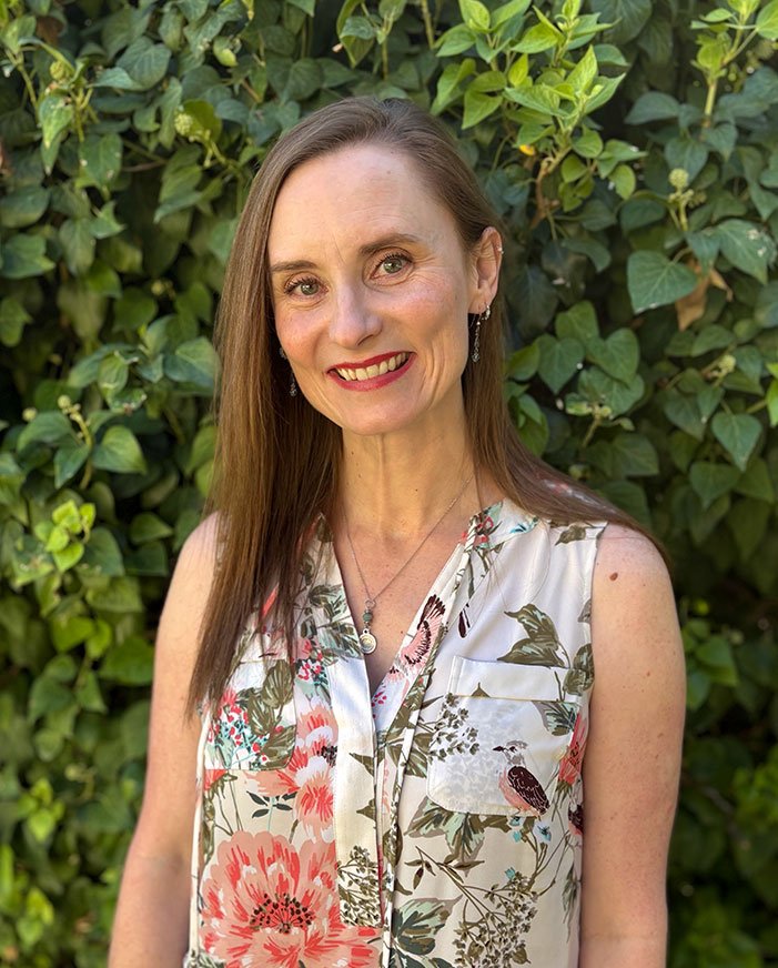 A woman with long brown hair wearing a sleeveless floral blouse, earrings, and a necklace, standing outdoors in front of green leafy plants.