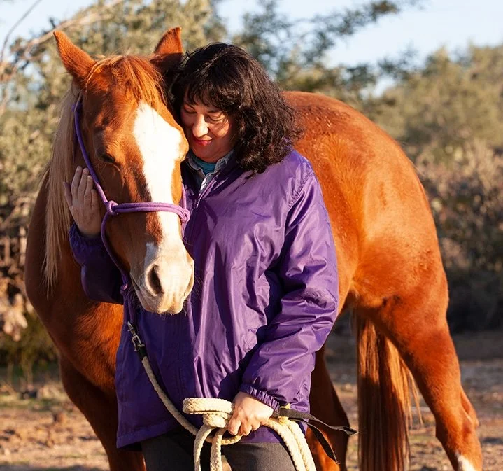 Woman with curly dark hair hugging a chestnut horse with a white blaze on its face, outdoors during daytime.