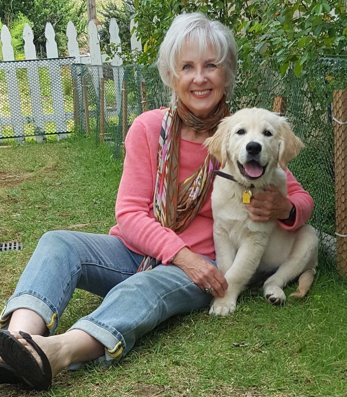 An elderly woman sitting on the grass outdoors, smiling, with a golden retriever puppy. She is wearing a pink top, blue jeans, and a striped scarf, with a brown bracelet. The puppy is sitting beside her, happy, with its tongue out, wearing a collar with a yellow tag.