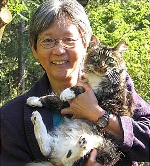 An older woman with glasses holding a large, long-haired tabby cat outdoors with greenery in the background.