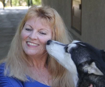 A woman with long blonde hair smiling as a black-and-white husky licks her face.