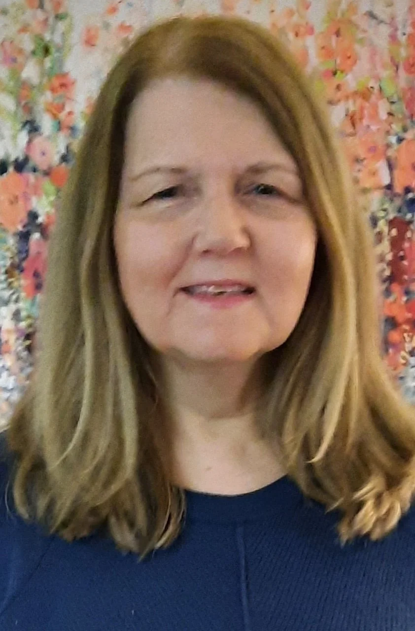 A woman with shoulder-length light brown hair and light skin, smiling slightly, standing in front of a colorful, floral-patterned background.