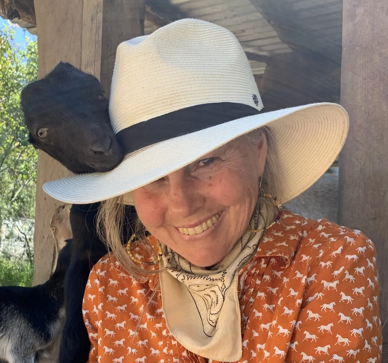 A woman wearing a large white sunhat with a black band, an orange shirt with white horse patterns, and a beige bandana around her neck, smiling outdoors with goats in the background.