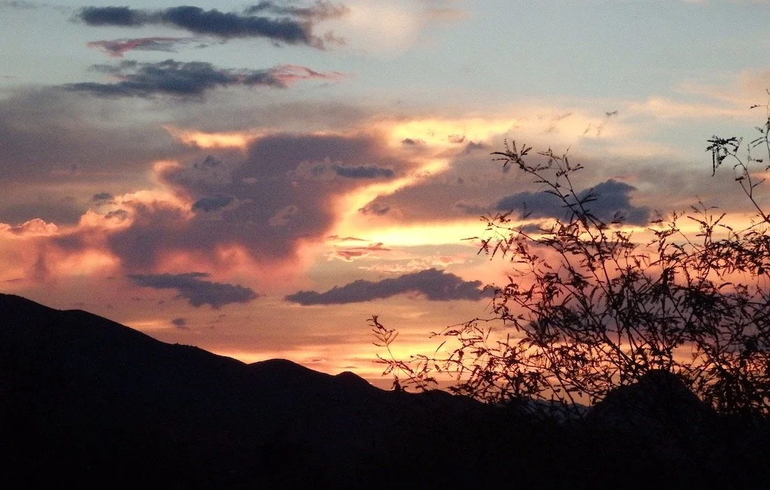 Sunset over silhouetted mountains with a tree in the foreground and colorful clouds in the sky.