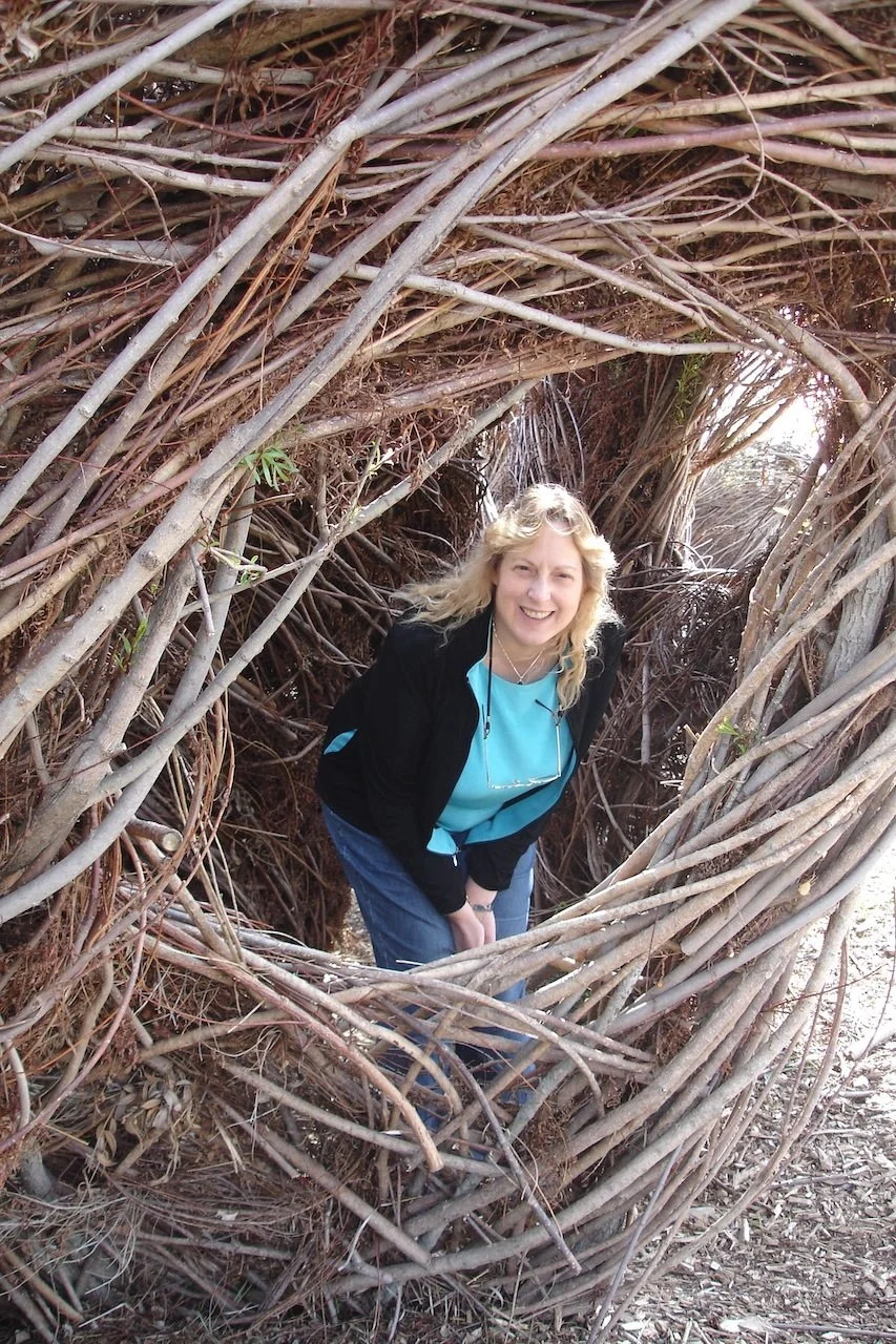 A woman with blonde hair, smiling, standing inside a large nest made of intertwined branches and twigs.