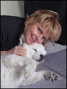 A woman with blonde hair lying down, hugging a white puppy with black eyes and nose, both lying on a gray blanket.