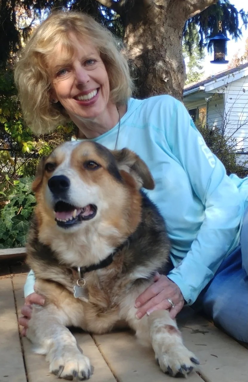 A woman with blonde, curly hair smiling and sitting on the ground next to a, medium-sized, mixed breed dog with a tricolor coat. They are outdoors in a yard with trees, bushes, and a house in the background.