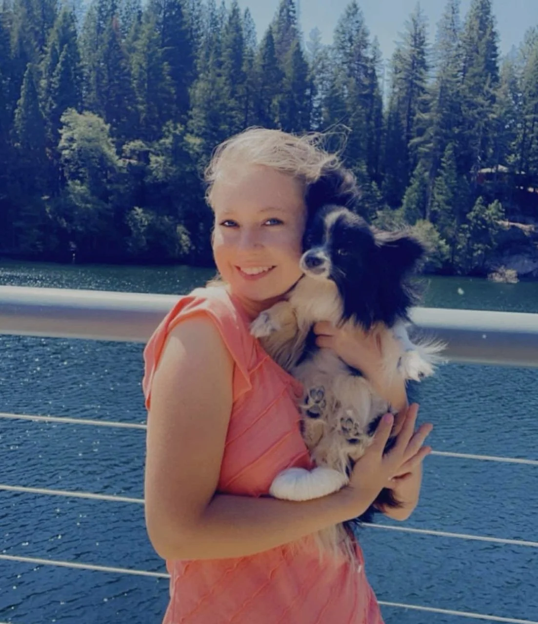 A young girl with blonde hair in a pink dress holding a black and white fluffy dog on a boat, with a lake and forested mountains in the background.