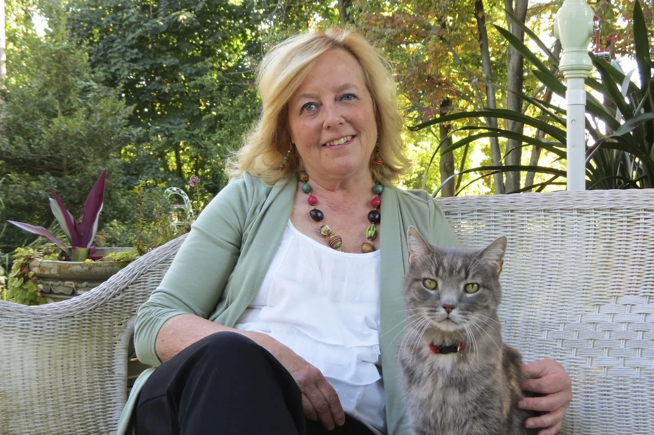 A woman sitting on a white outdoor wicker sofa with a gray cat on her lap, in a garden with green trees and plants in the background.