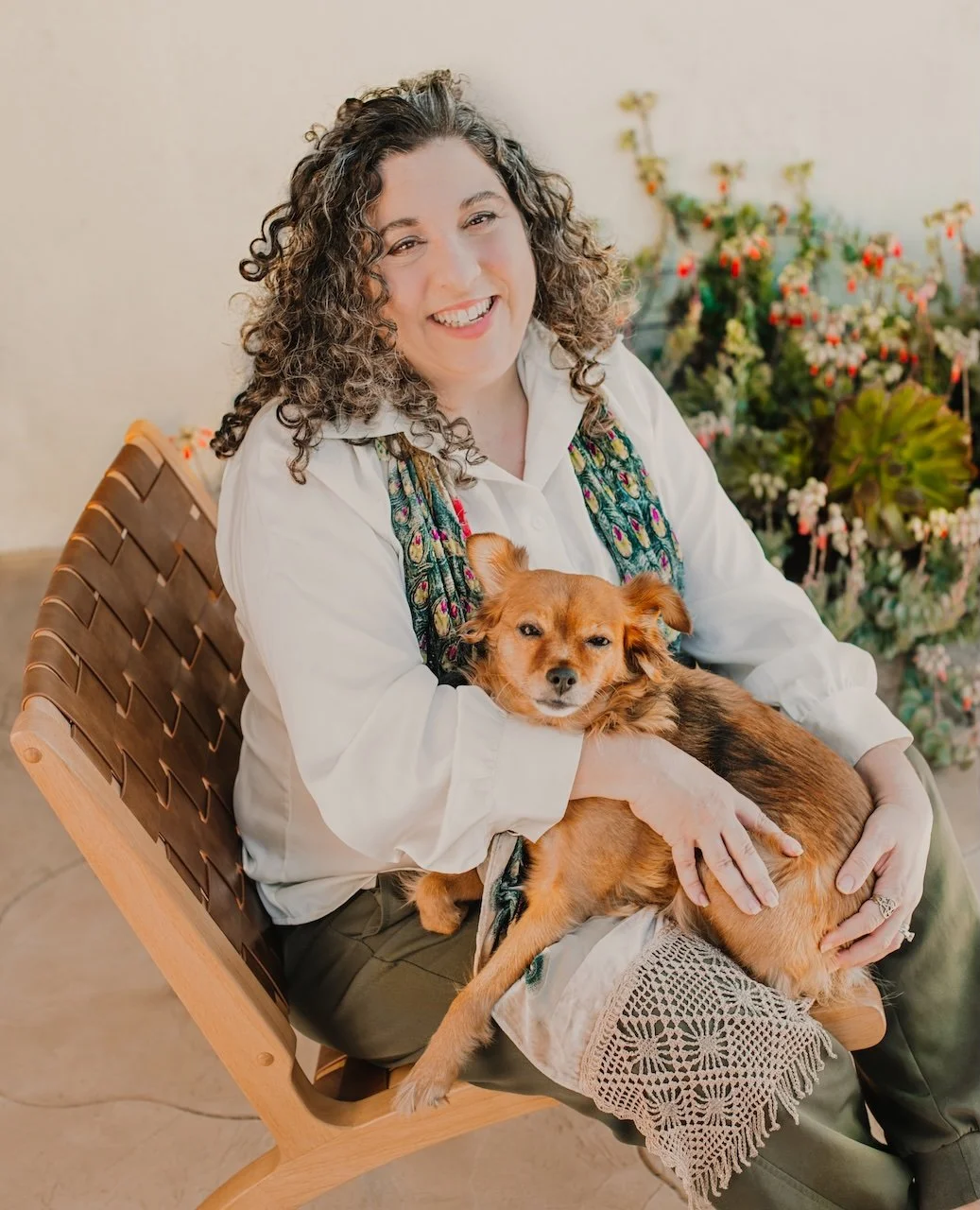 A woman with curly hair smiling and holding a small brown dog with long ears, sitting outdoors on a wooden chair with plants in the background.