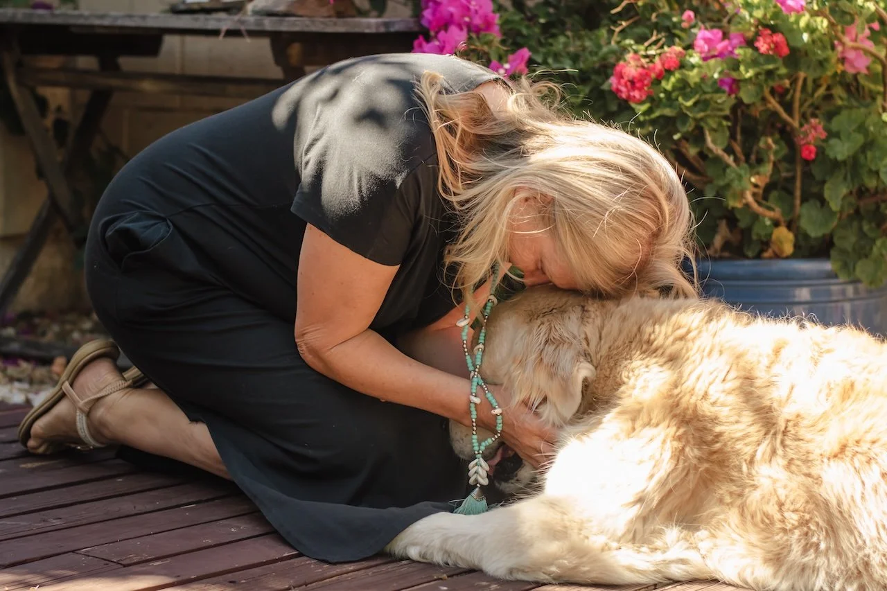 A woman kneeling on a wooden deck, hugging and kissing a large fluffy dog, with pink and purple flowers in a pot in the background.