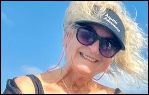 Smiling woman with blonde curly hair wearing sunglasses, a SunProtect visor, and a black top, outdoors on a sunny day.