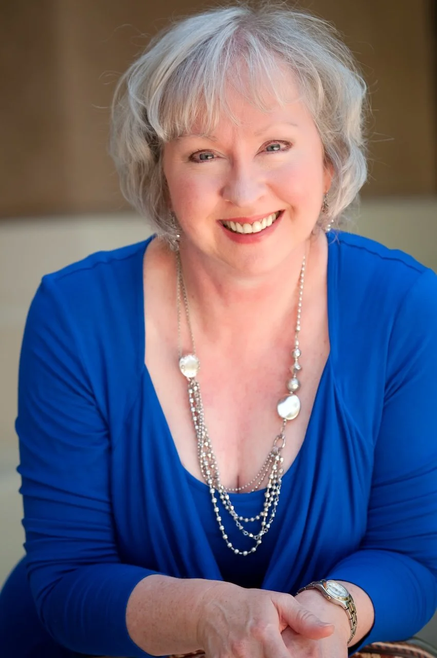 A smiling middle-aged woman with short gray hair, wearing a blue top, layered pearl necklaces, and a wristwatch, posed indoors against a blurred neutral background.
