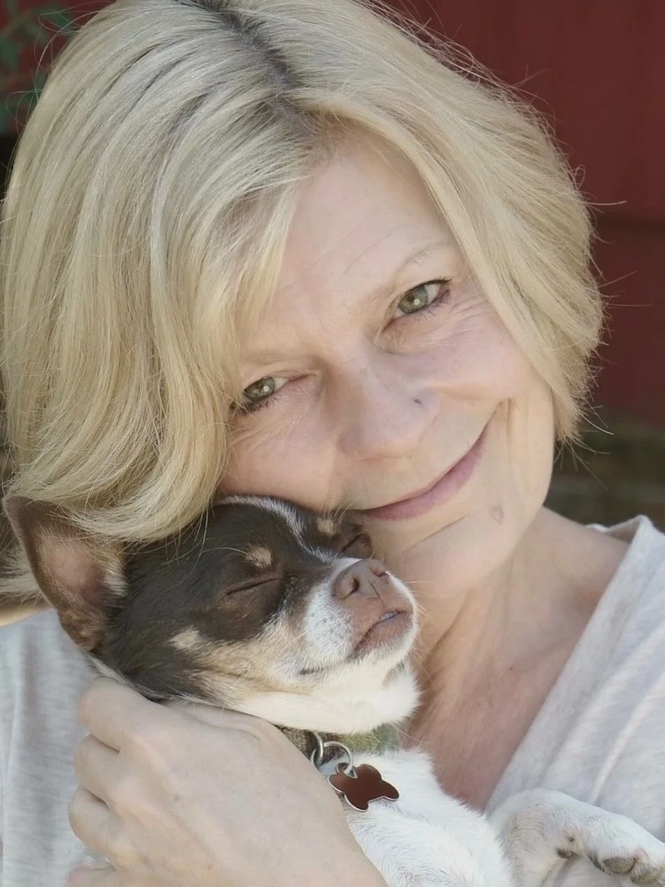 An elderly woman with short blonde hair smiling and holding a small black and white puppy close to her face, both appearing content and relaxed.