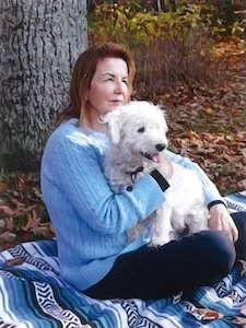 A woman sitting outdoors against a tree, holding a white fluffy dog. Fall leaves are visible in the background.