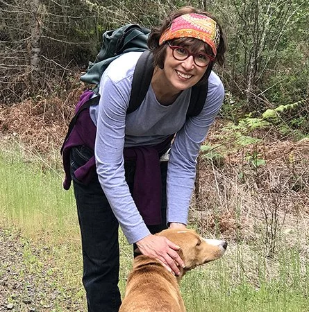 A woman smiling outdoors on a hike, with a backpack and a dog, standing on a trail surrounded by trees and greenery.