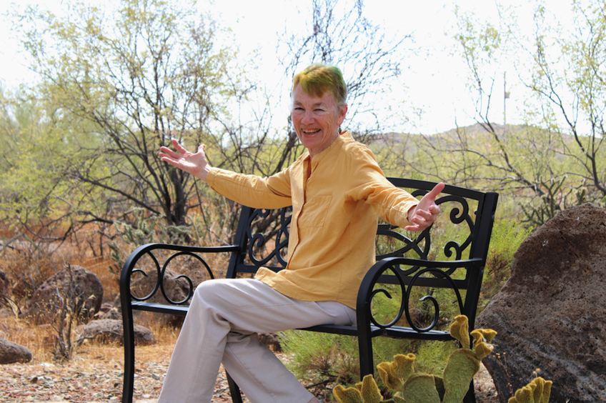 A smiling older woman sitting on a black decorative bench outdoors, with desert plants and rocks around her, and trees in the background.