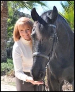 A young girl with blonde hair smiling and holding a black horse's chin outside, with trees and tropical plants in the background.