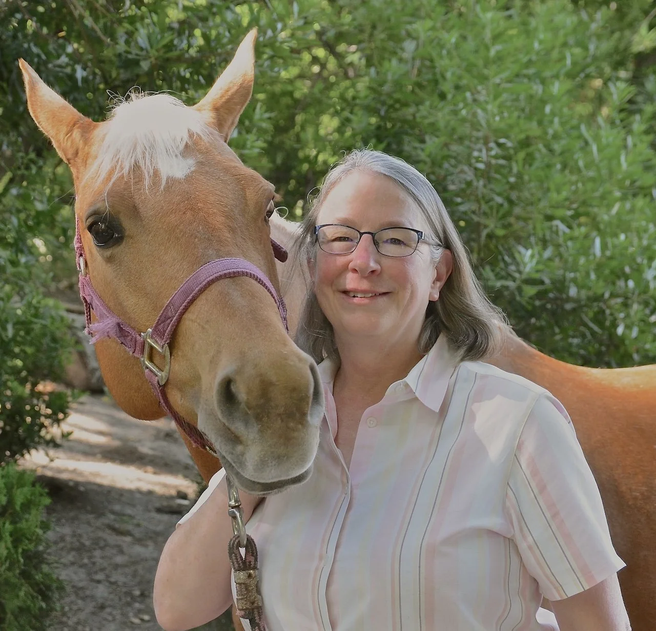 A woman with glasses and gray hair smiling next to a light brown horse with a pink halter in a green outdoor setting.