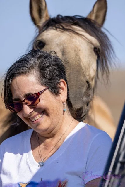 A woman smiling and wearing sunglasses outdoors with a horse behind her.