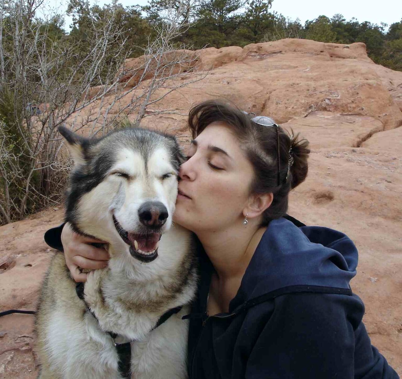 A woman and a Siberian husky dog outdoors on rocky terrain, with the woman kissing the dog on the cheek.