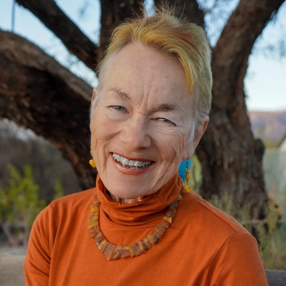 Close-up of an elderly woman smiling outdoors, wearing an orange top and jewelry, with a tree and natural landscape in the background.