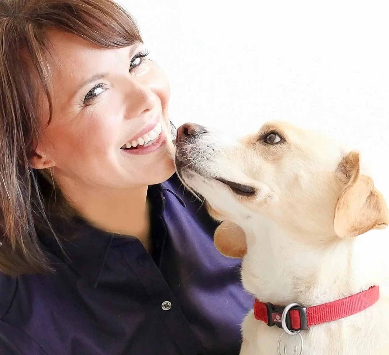 Woman smiling with a yellow labrador retriever puppy sniffing her face, both against a white background.