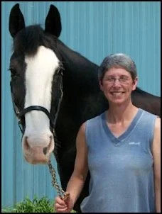 A woman smiling and holding a lead rope stand next to a black and white horse in front of a blue fence.