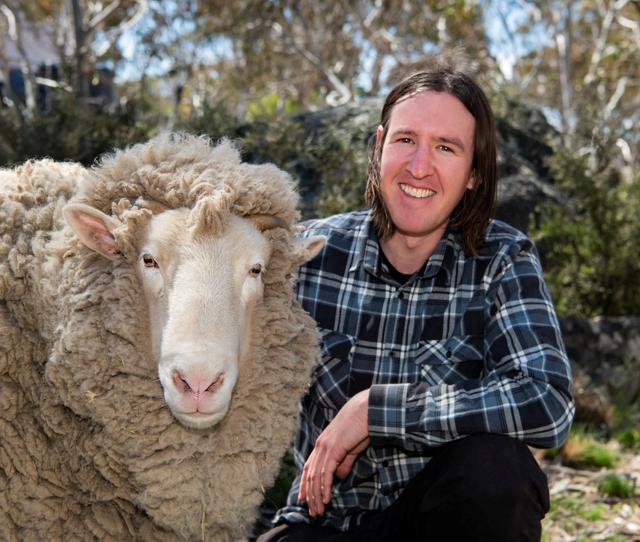 A man in a plaid shirt smiling next to a sheep with a thick, curly wool coat outdoors in a natural setting.