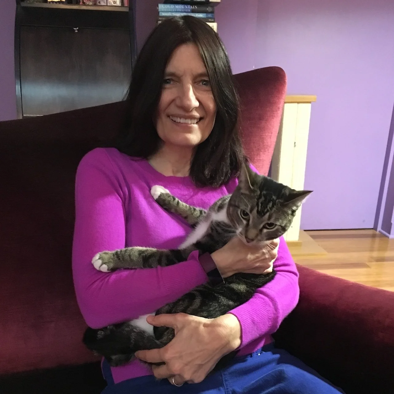 A woman with dark hair wearing a purple sweater sitting on a sofa, holding a tabby cat with stripes and white paws.
