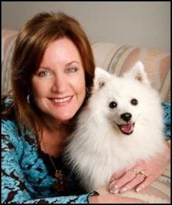 A woman smiling and holding a white fluffy dog indoors, with a bed in the background.