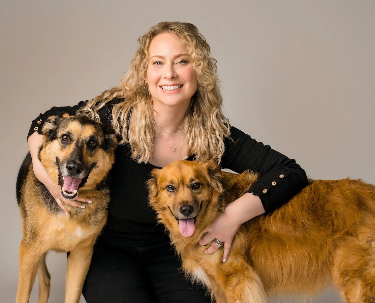 A woman with blonde curly hair smiling and posing with two Golden Retrievers against a plain gray background.