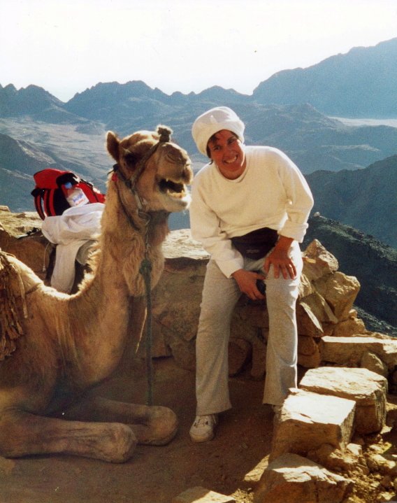 A woman smiling and crouching next to a camel on a rocky mountain trail with a scenic mountain range in the background.