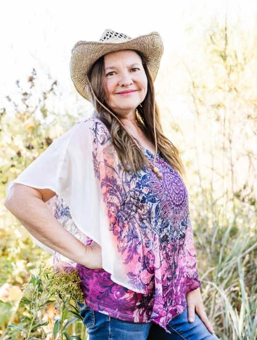 A woman wearing a straw hat and a colorful paisley top, standing outdoors in a natural setting with trees and plants, smiling at the camera.