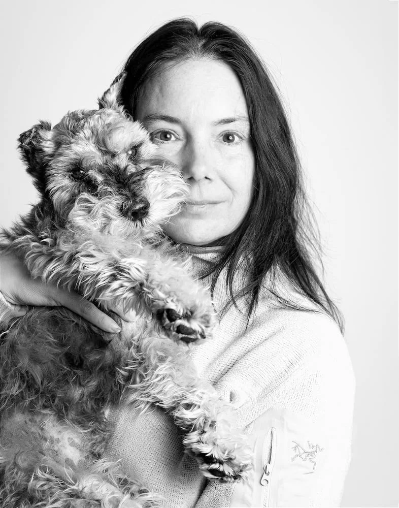 A woman with long dark hair holding a fluffy, small dog with curly fur close to her face.