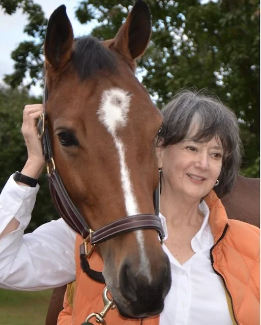 A woman with gray hair smiling next to a brown horse with a white stripe on its face in a green outdoor setting.