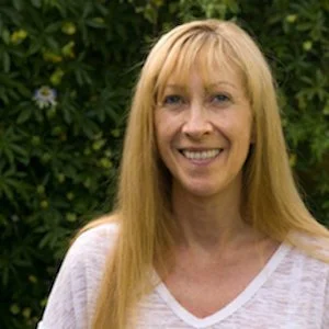 A woman with long blonde hair smiling outdoors in front of green foliage.