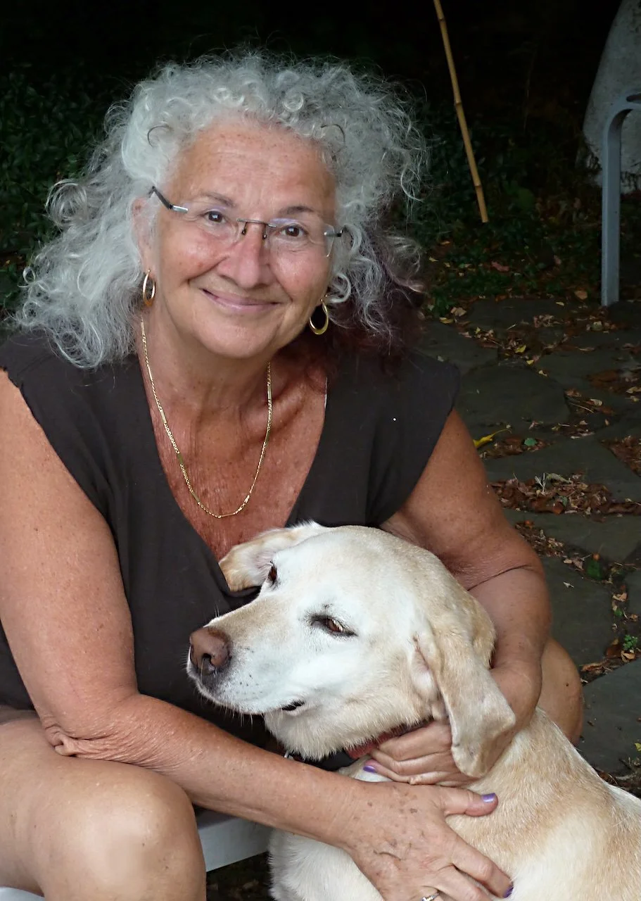An elderly woman with curly gray hair, glasses, and gold jewelry is smiling while holding a light-colored Labrador Retriever dog outdoors on a stone pathway.