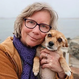 A woman with gray hair, wearing glasses, a purple scarf, and a brown jacket, holding a small brown and white dog outdoors near water.