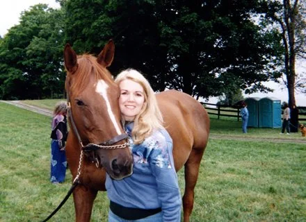 A woman with blonde hair standing next to a brown horse with a white stripe on its face in a grassy area with trees and people in the background.