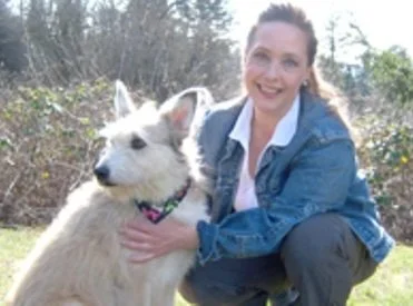 A woman with brown hair and a blue jacket smiling while kneeling next to a large, fluffy white dog outdoors.