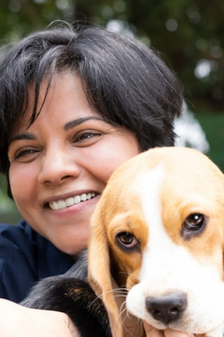 A woman smiling and hugging a beagle puppy with a blurred outdoor background.
