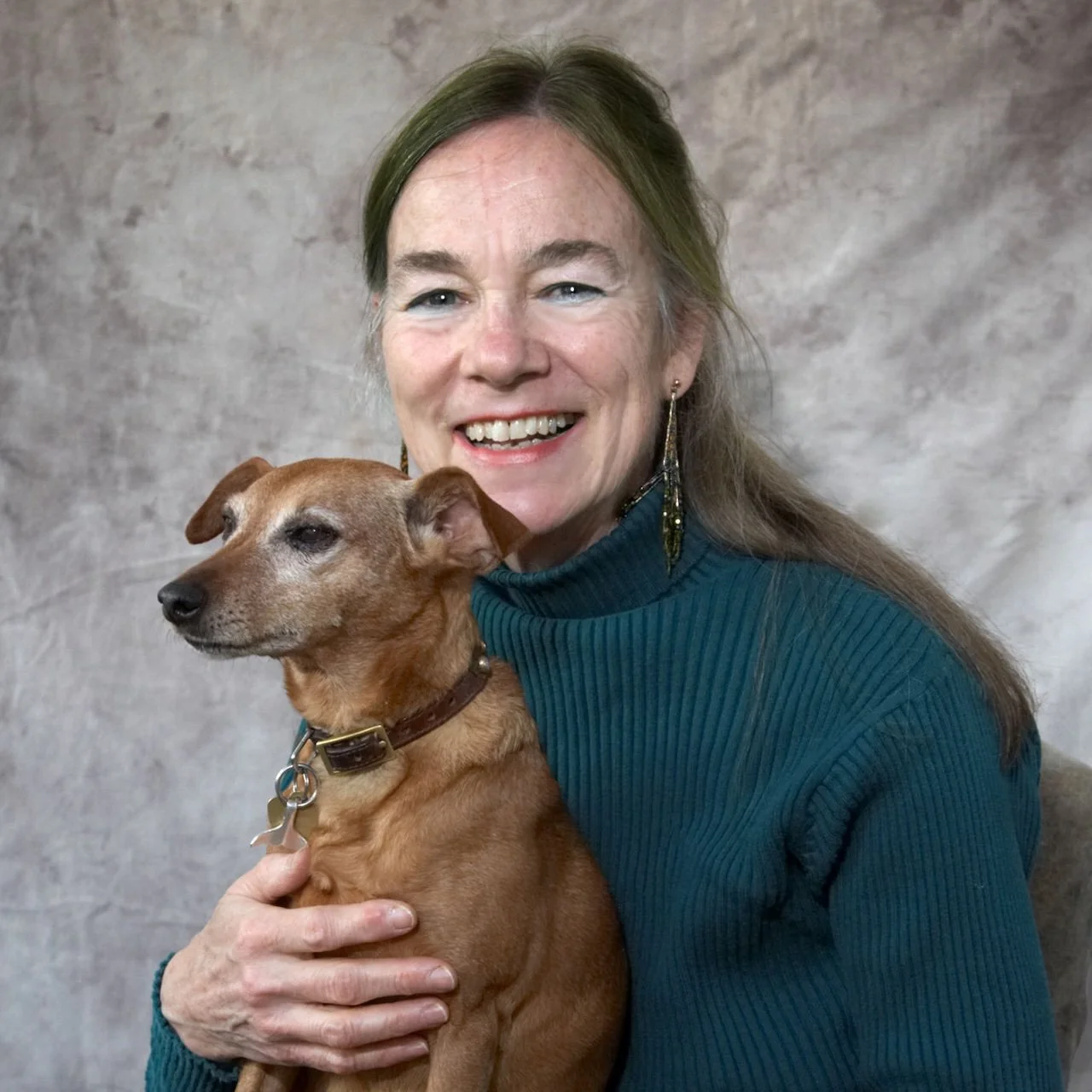 A smiling woman with long, light brown hair wearing a teal ribbed turtleneck holds a small brown dog with a collars and a nose that looks like it is either black or dark brown. The woman is wearing long earrings and standing against a plain gray background.