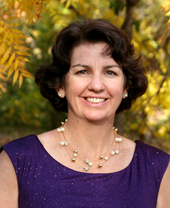 A woman with short, dark curly hair smiling outdoors with yellow and green autumn leaves in the background, wearing a purple sleeveless dress and pearl jewelry.