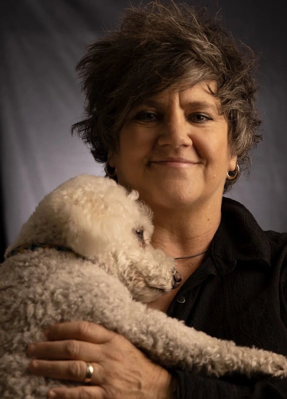 A woman with short, curly hair and wearing earrings is smiling while holding a small, fluffy white dog close to her chest.