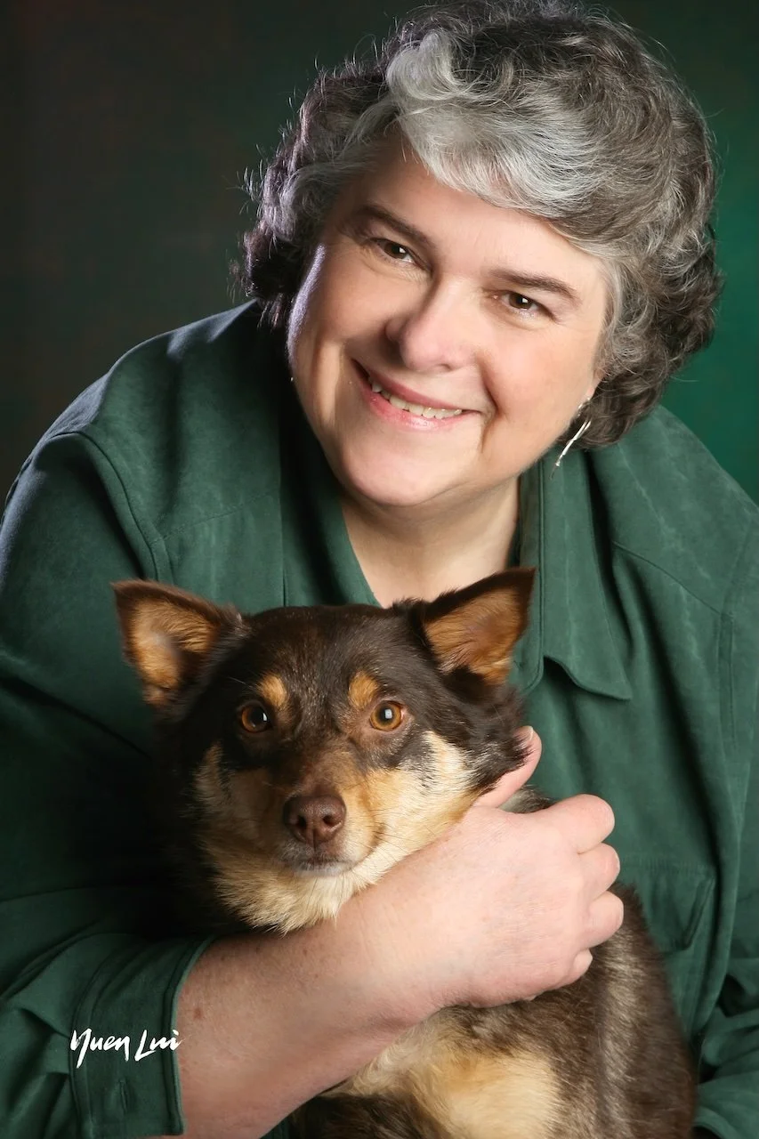 A woman with gray curly hair smiling and holding a small brown and tan dog.