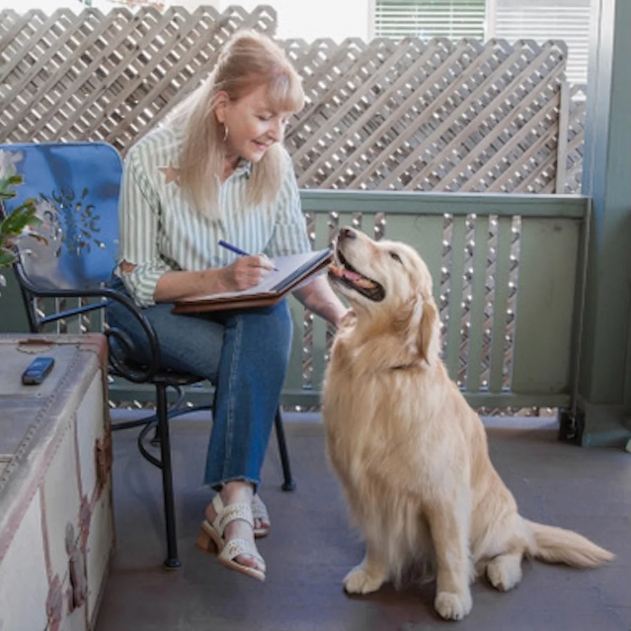 A woman with blonde hair in a striped shirt sitting on a chair, writing in a notebook, with a golden retriever sitting next to her on a porch.