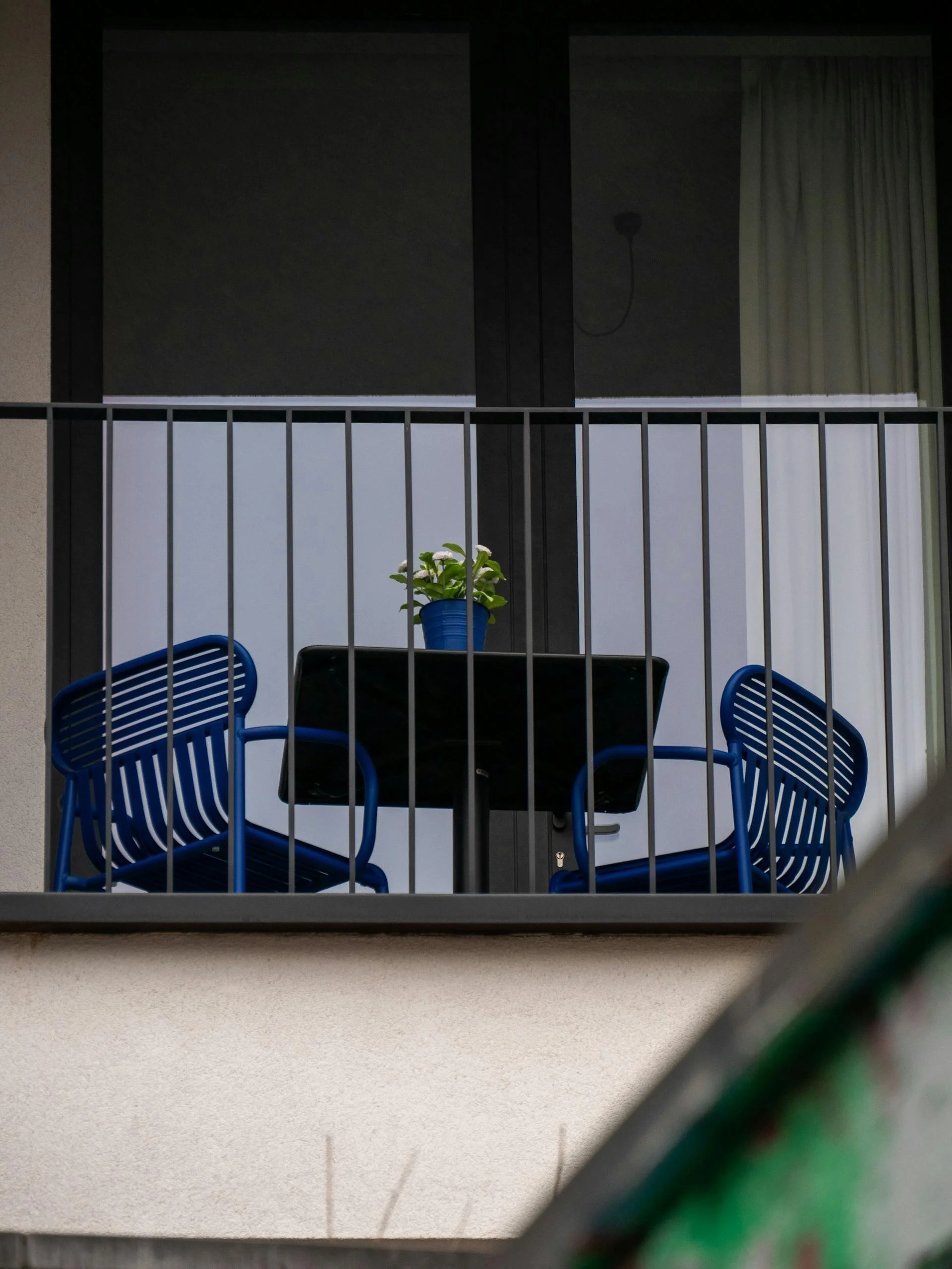 A balcony with a black table, four blue chairs, a potted plant with white flowers, and a background of sliding glass doors with curtains, viewed through a black railing.