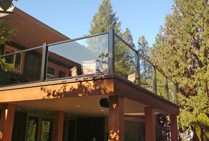 Backyard deck with glass railing, part of a wooden house, surrounded by trees and sunlight.