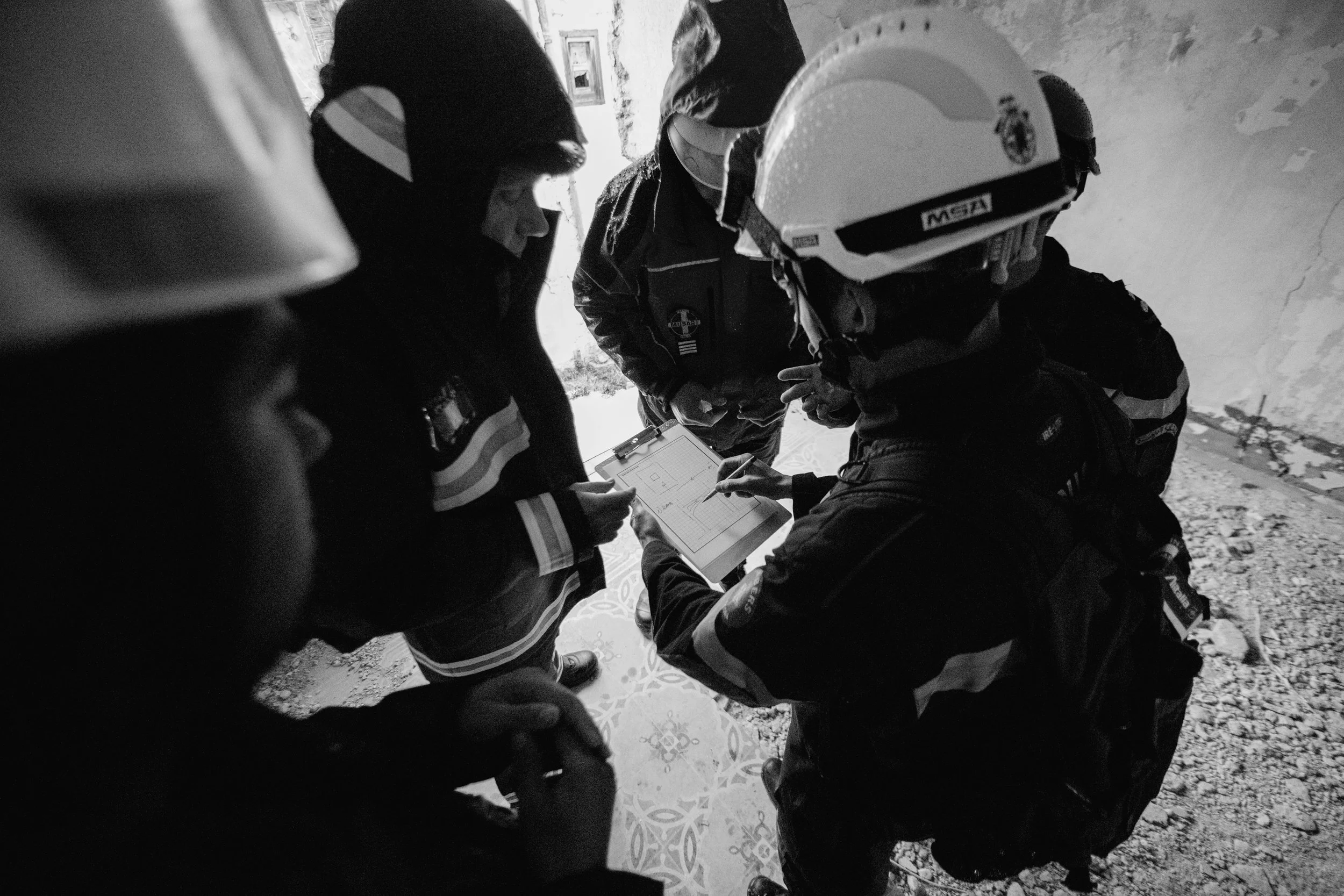 A group of individuals, wearing helmets and jackets, gathered around a clipboard or tablet, possibly discussing or reviewing plans or instructions, outdoors near a mountainous area.